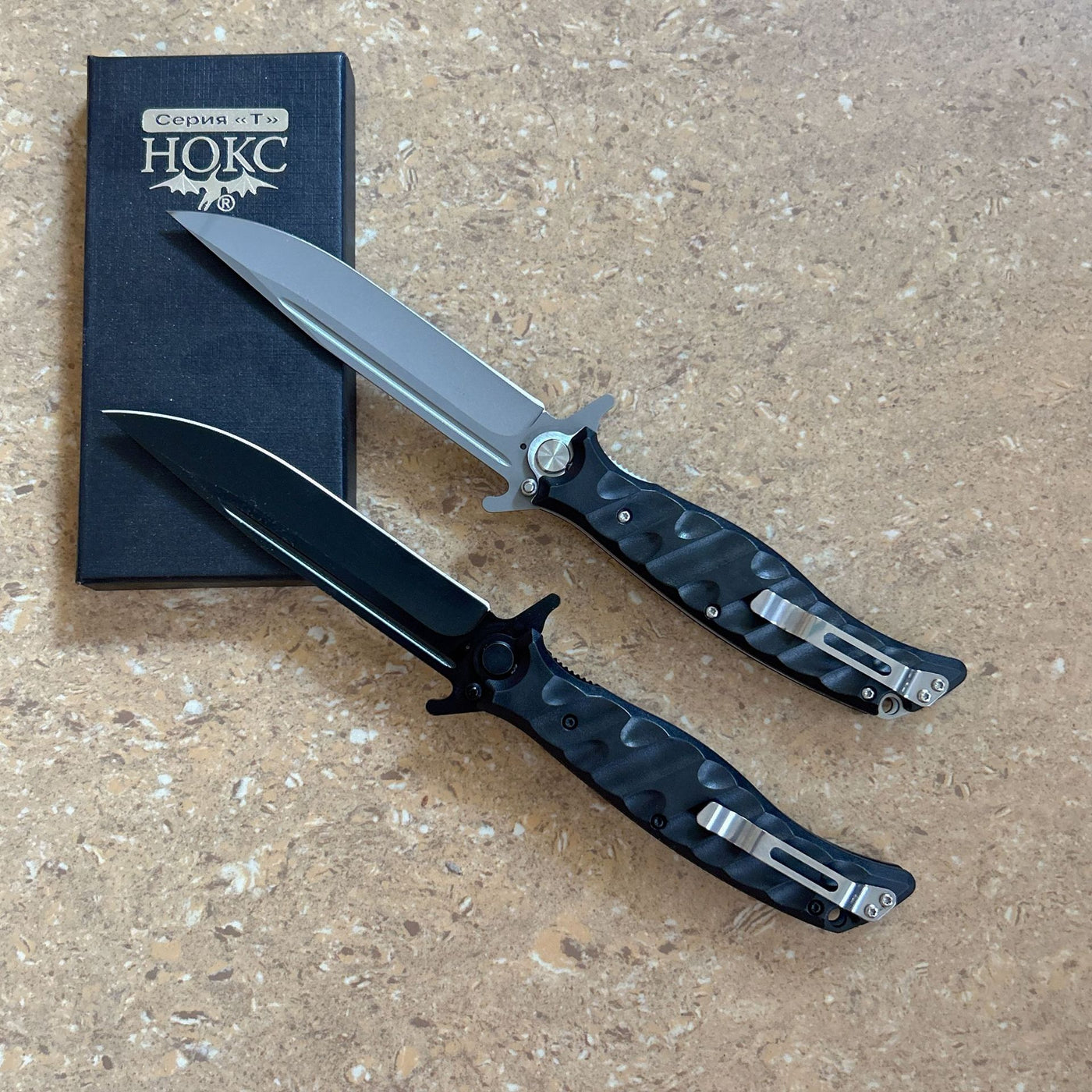 Two Noks folding knives with a black box on a light wooden surface. The top knife has a silver blade, while the bottom knife has a black blade.