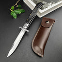 An open Italian stiletto knife with a black handle and silver blade, displayed next to a brown leather sheath on a dark textured surface.
