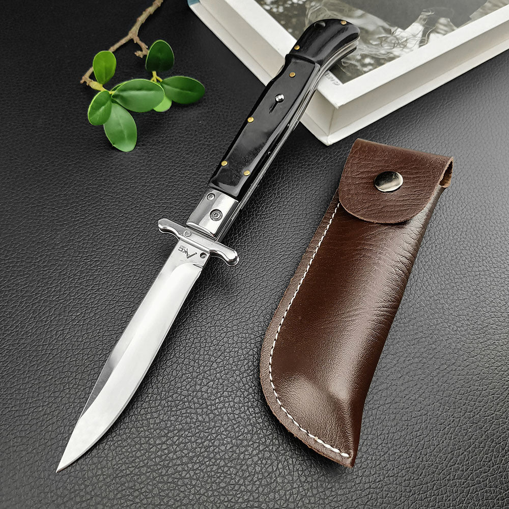 An open Italian stiletto knife with a black handle and silver blade, displayed next to a brown leather sheath on a dark textured surface.