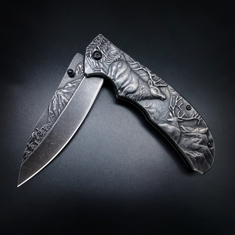An open folding knife featuring detailed relief engravings of deer and mountain forest landscapes on the handle and blade, displayed on a dark textured background.