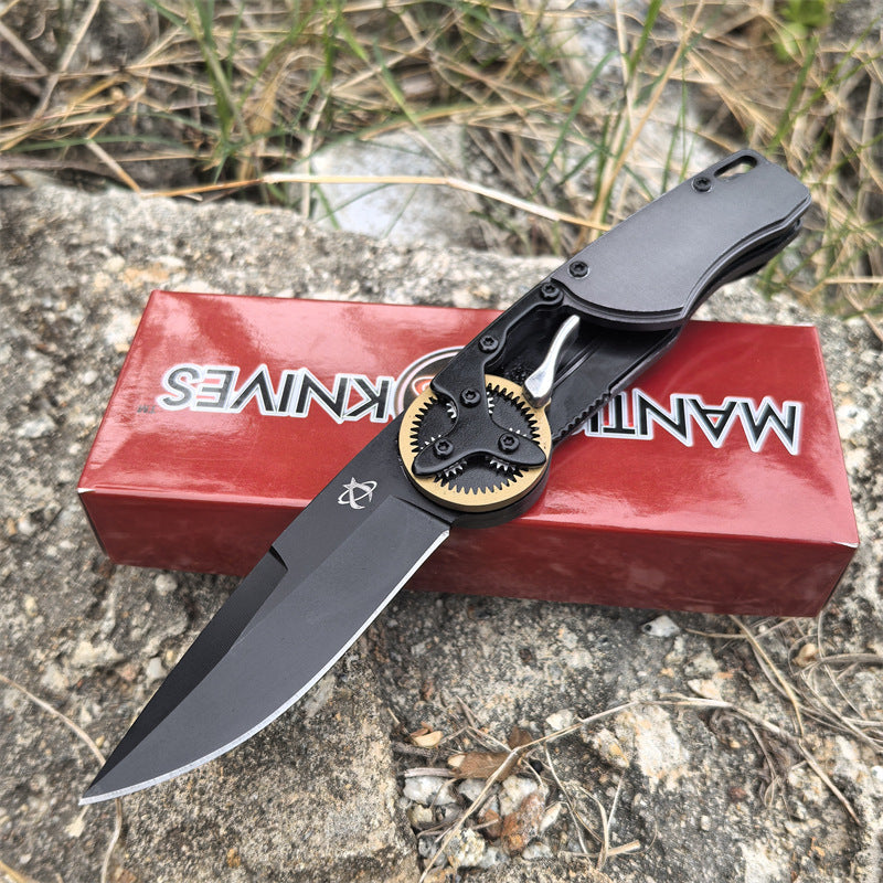 An open folding knife with a black blade and gold gear pivot, displayed resting on a red packaging box outdoors.