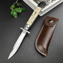 An open Italian stiletto knife with a cream bone handle and silver blade, displayed next to a brown leather sheath on a dark textured surface.
