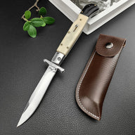 An open Italian stiletto knife with a cream bone handle and silver blade, displayed next to a brown leather sheath on a dark textured surface.