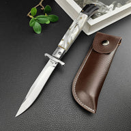 An open Italian stiletto knife with a white pearl handle and silver blade, displayed next to a brown leather sheath with white stitching on a dark textured surface.