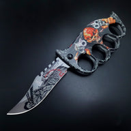 An open knuckle duster folding knife with a black and orange skull graphic handle and a partially serrated silver blade, displayed on a dark background.