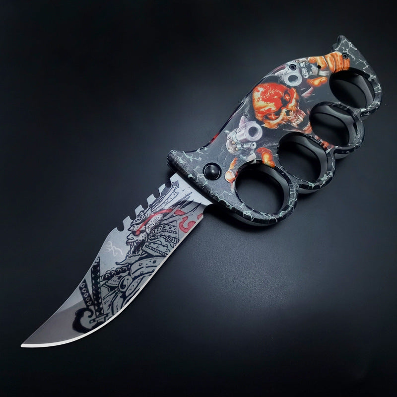 An open knuckle duster folding knife with a black and orange skull graphic handle and a partially serrated silver blade, displayed on a dark background.