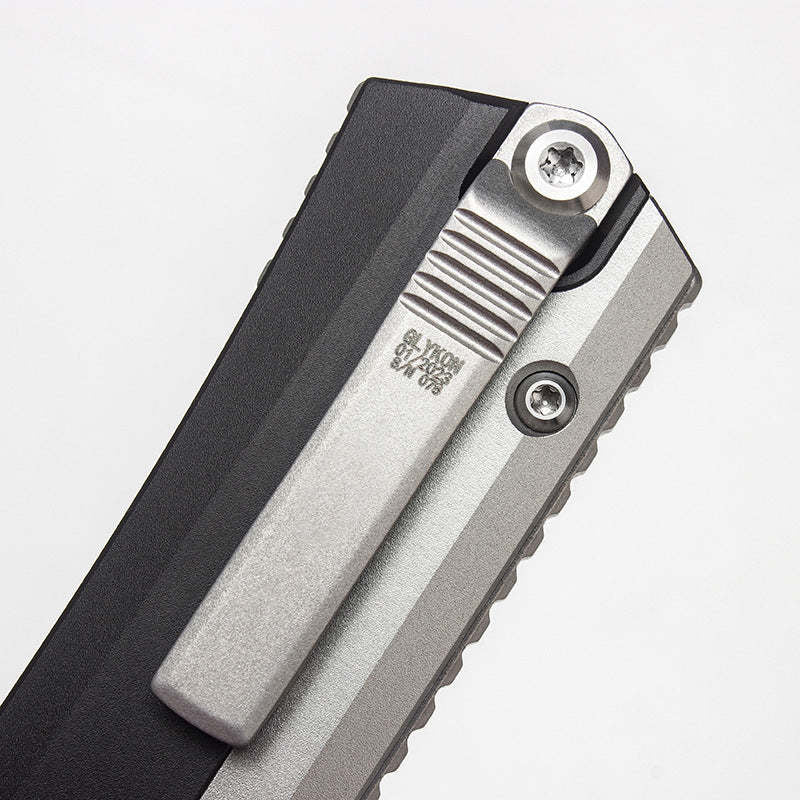 A close-up of a silver pocket clip on a black out-the-front knife, showing engraved text and serial numbers on a white background.