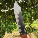 Alt Text: A close-up of a fixed blade knife with a hammered blade and wooden handle held in front of green tree leaves outdoors.
