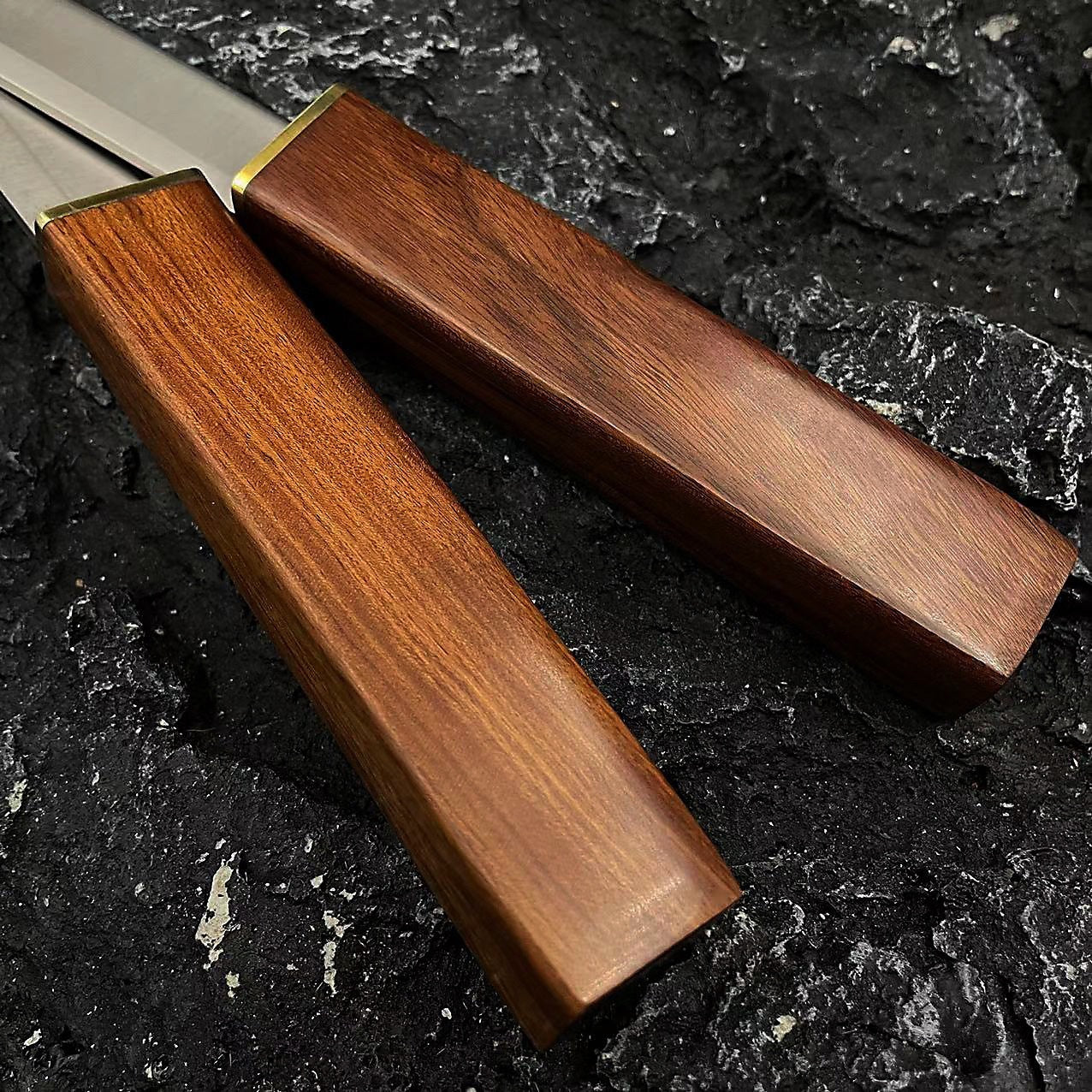 Close-up of two knife handles featuring polished wood with a smooth finish and brass guards, resting on a dark, rough rock background.