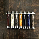 A collection of seven closed stiletto folding knives with a variety of handle styles including wood, white pearl, snakeskin, orange resin, black, and blue resin, lined up on a rustic wooden background.