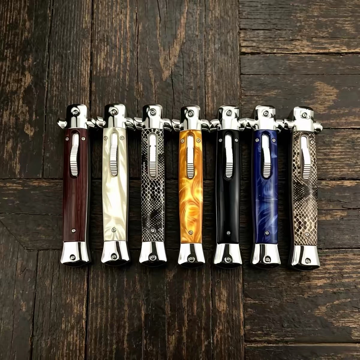 A collection of seven closed stiletto folding knives with a variety of handle styles including wood, white pearl, snakeskin, orange resin, black, and blue resin, lined up on a rustic wooden background.