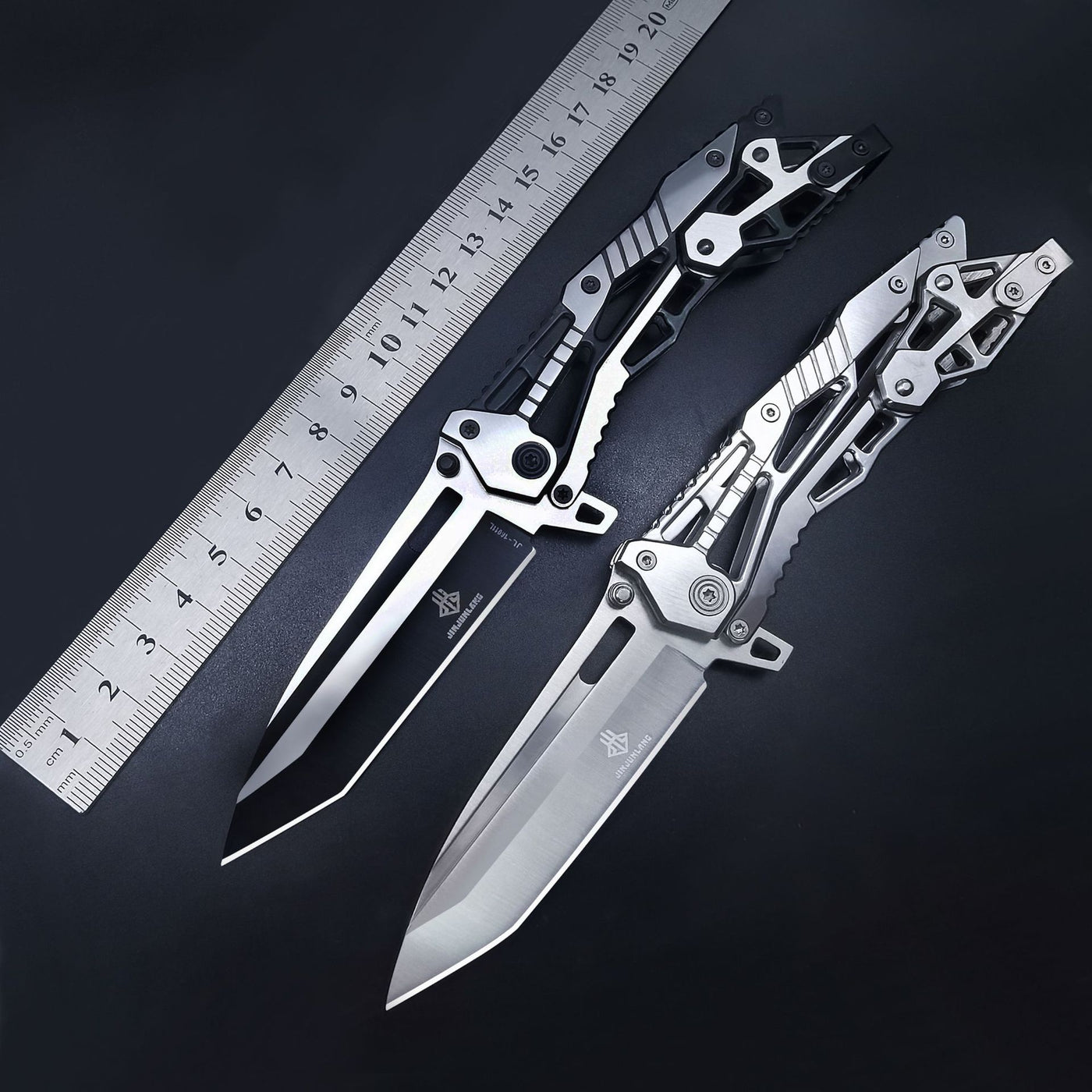 Two open skeletonized Tanto folding knives, one black and one silver, placed next to a metal ruler on a dark background to show their size and length.