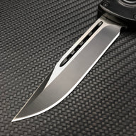A close-up of a silver folding knife blade showing a central groove and a brushed metal finish, displayed on a textured dark background.