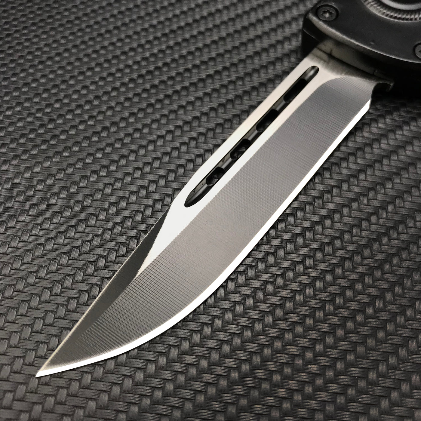 A close-up of a silver folding knife blade showing a central groove and a brushed metal finish, displayed on a textured dark background.