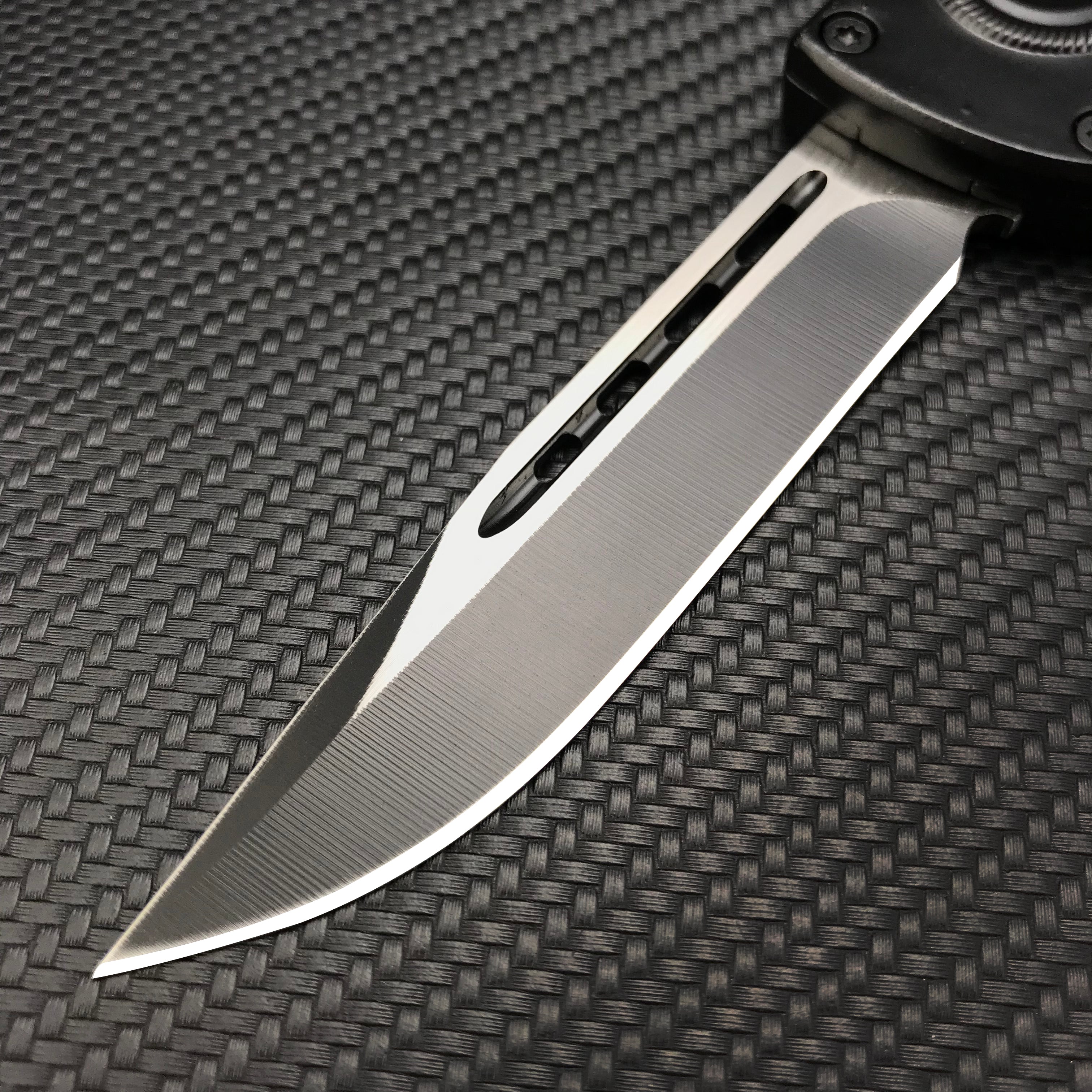 A close-up of a silver folding knife blade showing a central groove and a brushed metal finish, displayed on a textured dark background.