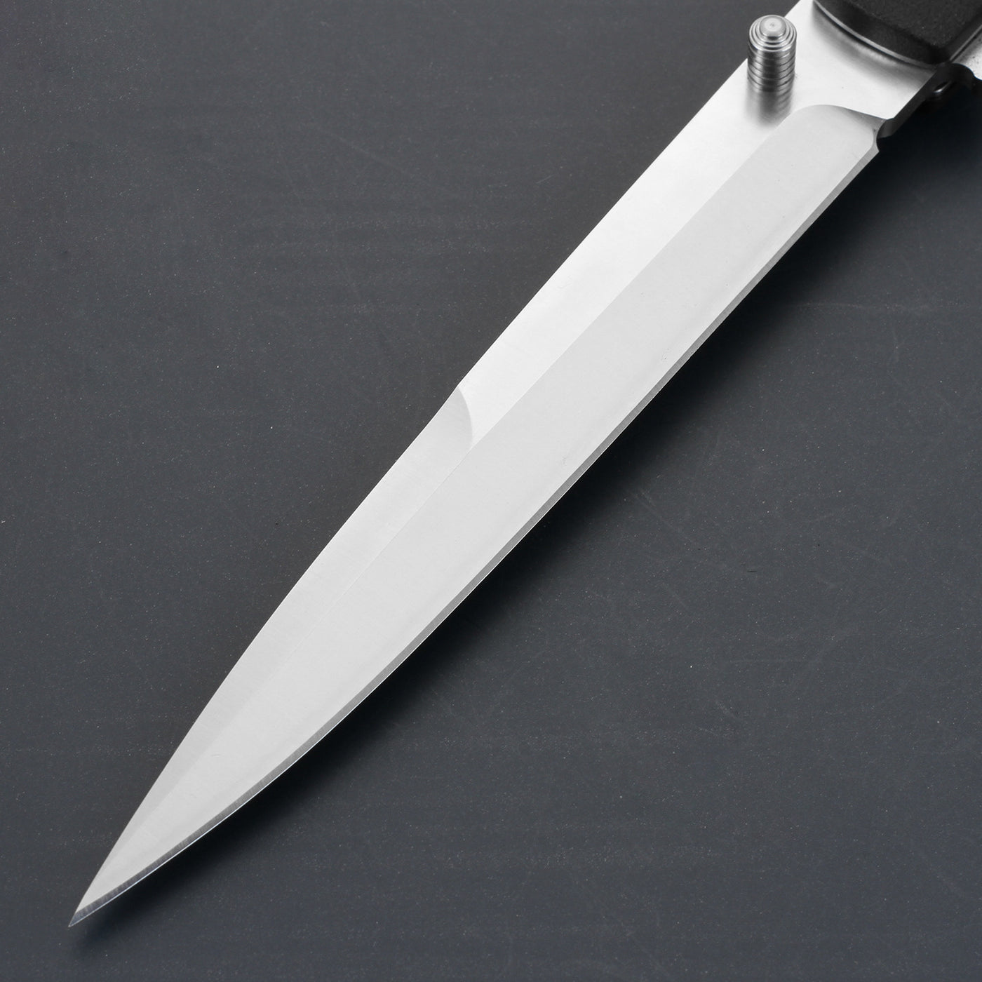 A close-up of a sharp silver stainless steel folding knife blade with a cylindrical thumb stud, displayed on a dark grey surface.