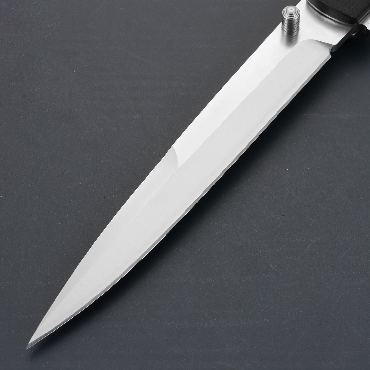 A close-up of a sharp silver stainless steel folding knife blade with a cylindrical thumb stud, displayed on a dark grey surface.