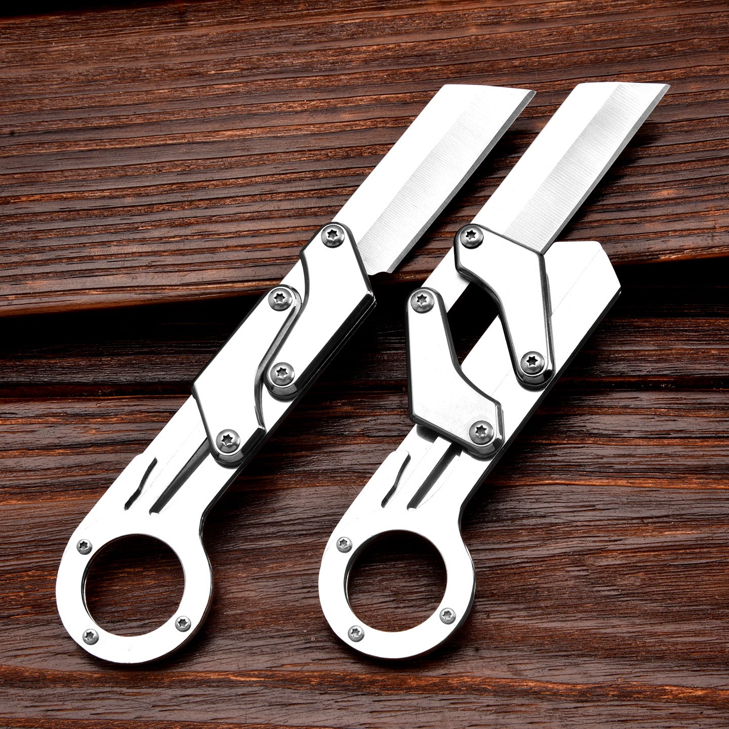 Two silver mechanical folding knives with ring handles and grooved blades, one fully opened and one partially folded to show the linkage mechanism, displayed on a dark wooden background.