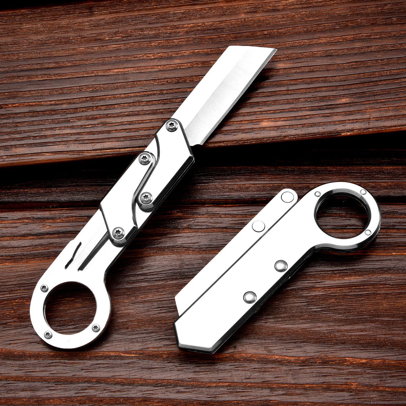Two silver folding knives with ring handles and mechanical linkage designs, one open and one closed, displayed on a dark wooden background.
