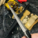 A handheld close-up of a slim tactical folding knife with a silver dagger blade, displayed against a rough dark background with a gold toad statue and red flower.
