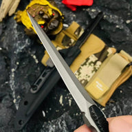 A handheld close-up of a slim tactical folding knife with a silver dagger blade, displayed against a rough dark background with a gold toad statue and red flower.
