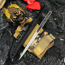 A tactical flat lay composition featuring a silver slim folding knife, black sheath, camo pouch, decorative gold toad statue, and a red flower on a dark stone surface.