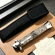 A closed stiletto knife with a silver and black snakeskin patterned handle, resting on a leather notebook next to a black nylon sheath and technical sketches.