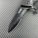 A close-up of the black serrated blade of a Tac-Force knuckle duster folding knife, showing the logo and full groove on a carbon fiber background.