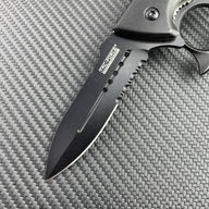 A close-up of the black serrated blade of a Tac-Force knuckle duster folding knife, showing the logo and full groove on a carbon fiber background.