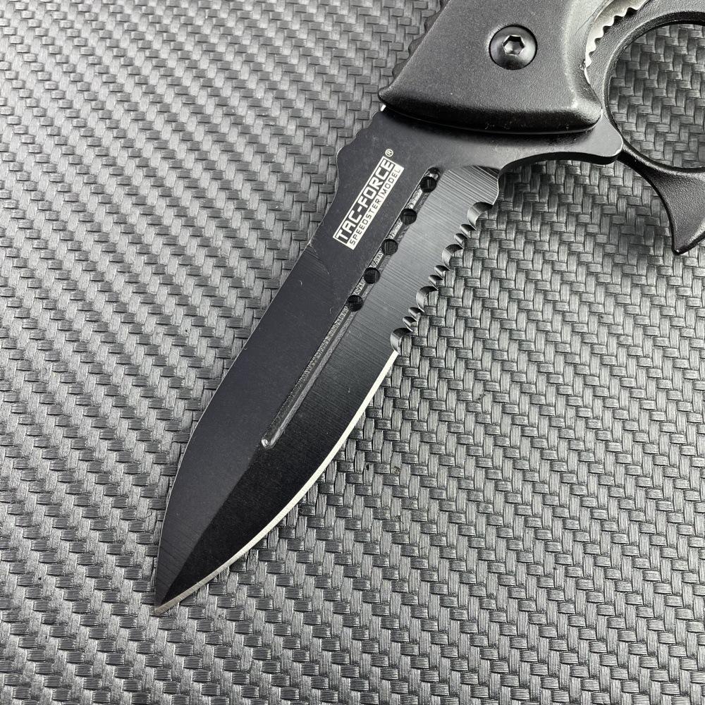 A close-up of the black serrated blade of a Tac-Force knuckle duster folding knife, showing the logo and full groove on a carbon fiber background.