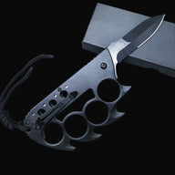 A closed black Tac-Force knuckle duster folding knife with a pocket clip and lanyard, resting on a black box against a dark background.