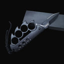 An open black Tac-Force knuckle duster folding knife with a partially serrated blade, silver logo, and black braided lanyard, displayed on a dark box against a dark background.
