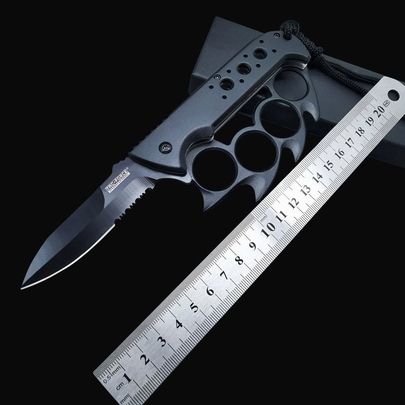 An open Tac-Force knuckle duster folding knife with a black partially serrated blade and lanyard, displayed next to a metal ruler on a dark background.