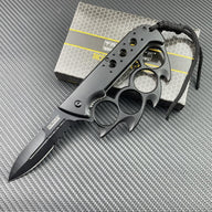 An open Tac-Force knuckle duster folding knife with a black serrated blade and lanyard, displayed next to its retail packaging box on a textured background.