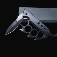 A partially open black Tac-Force knuckle duster folding knife with a black lanyard, resting on a black box against a dark background.
