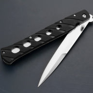 A close-up of an open Tac-Raven folding knife, showing a silver blade and a black handle with circular cutouts, resting on a dark surface.