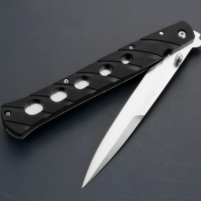 A close-up of an open Tac-Raven folding knife, showing a silver blade and a black handle with circular cutouts, resting on a dark surface.