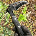 Black tactical multi-purpose claw knife/breaching tool held in hand, with green outdoor foliage in the background, showcasing its practical functionality and rugged tactical style.