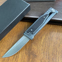 An open tactical folding knife with a drop point blade displayed next to its black packaging box on a wooden surface.