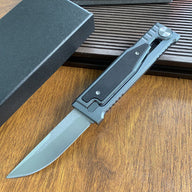 An open tactical folding knife with a drop point blade displayed next to its black packaging box on a wooden surface.