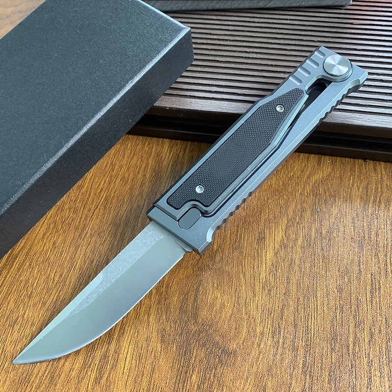 An open tactical folding knife with a drop point blade displayed next to its black packaging box on a wooden surface.