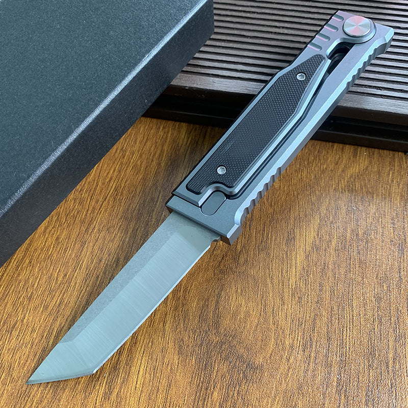 An open tactical folding knife with a tanto blade displayed next to its black packaging box on a wooden surface.