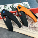 Two open tactical folding knives with black blades and geometric handles. The left knife is black with a blue pivot screw, and the right knife is orange. They are displayed diagonally on a red book featuring vintage black and white photos on a grey textured surface.