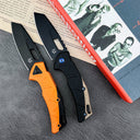 Two open tactical folding knives with black blades. The left knife has an orange handle, and the right knife features a black and tan handle with a blue pivot screw. They are displayed on a red "Trainspotting" book on a textured grey surface.