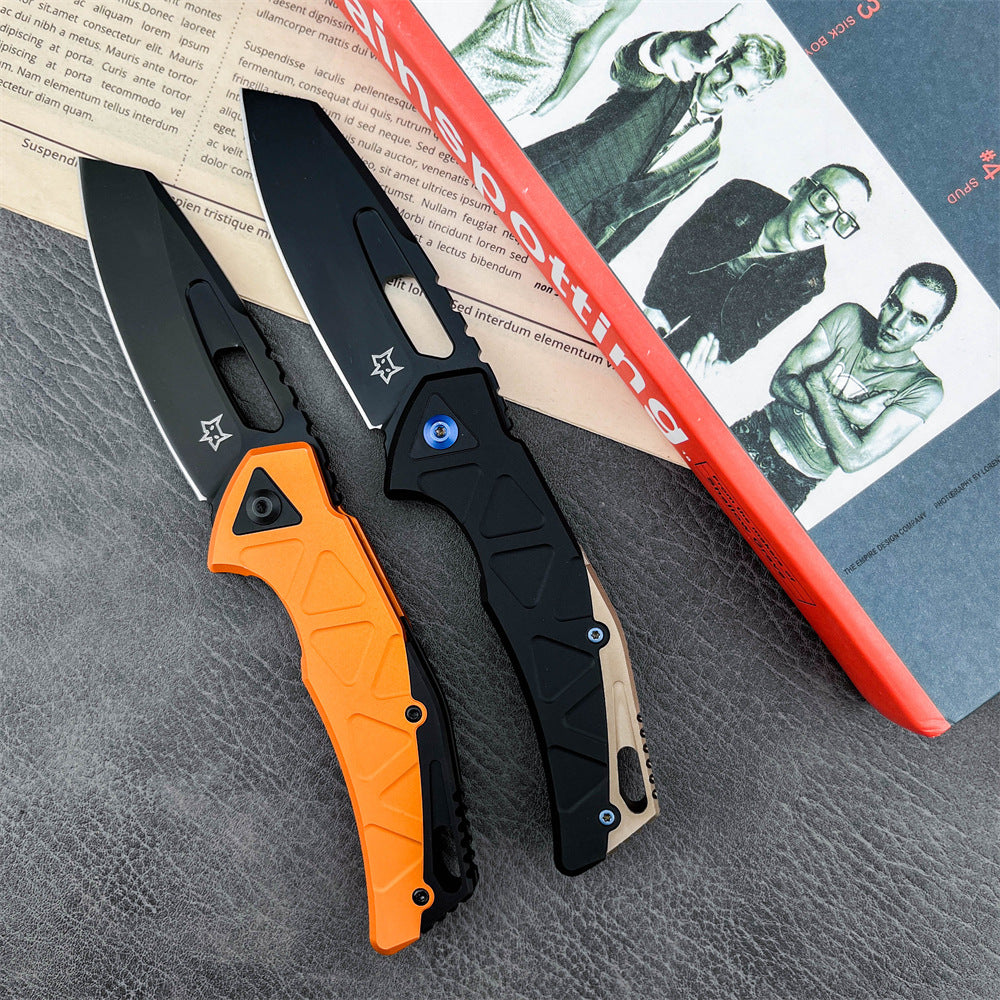 Two open tactical folding knives with black blades. The left knife has an orange handle, and the right knife features a black and tan handle with a blue pivot screw. They are displayed on a red "Trainspotting" book on a textured grey surface.