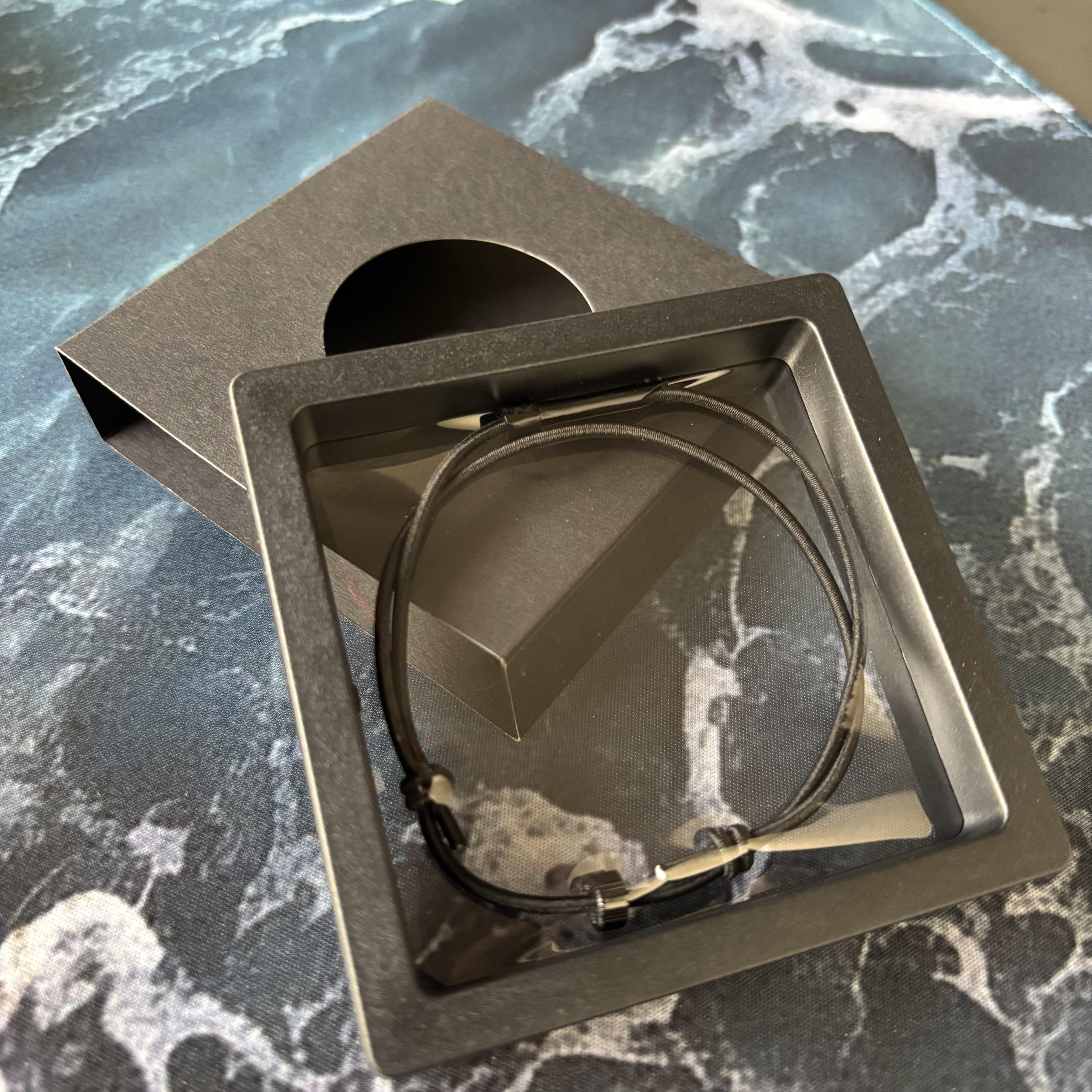 A black tactical handcuff key bracelet displayed inside a black presentation box with a clear window, resting on a blue marble-textured surface.
