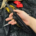A hand holding a slim tactical folding knife in its black sheath, showcasing the knife handle and the "ULTI CLIP" attachment on the sheath against a dark rocky background.