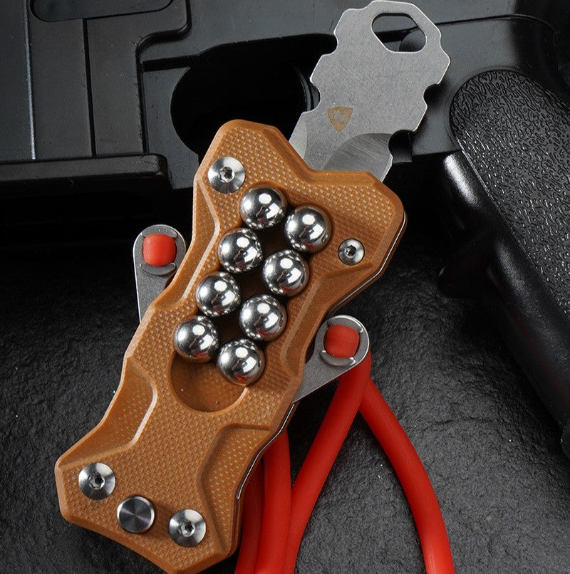 A tan G10 handle EDC multi-tool slingshot with a stainless steel fork and red rubber band, positioned with its 8Cr13MoV steel blade deployed against a dark tactical background.