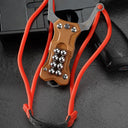 A tan G10 handle EDC slingshot with a red high-elastic band and steel ball storage, displayed with the band hung loosely on a dark tactical firearm background.
