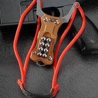 A tan G10 handle EDC slingshot with a red high-elastic band and steel ball storage, displayed with the band hung loosely on a dark tactical firearm background.